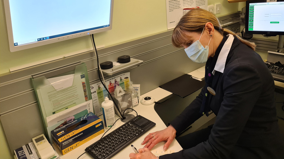 A woman with hair in a ponytail is leaning over a desk and writing with a pen in front of a mounted wall screen in an optometry testing room