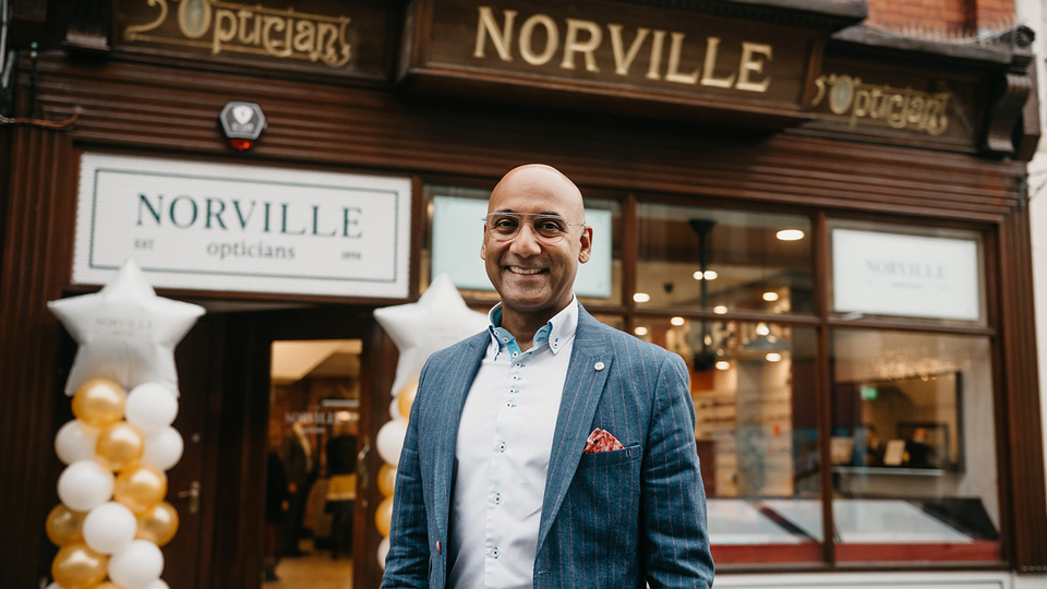 A man wearing glasses and a blue suit jacket is smiling in front of an optometry practice, which has a historic wooden frontage