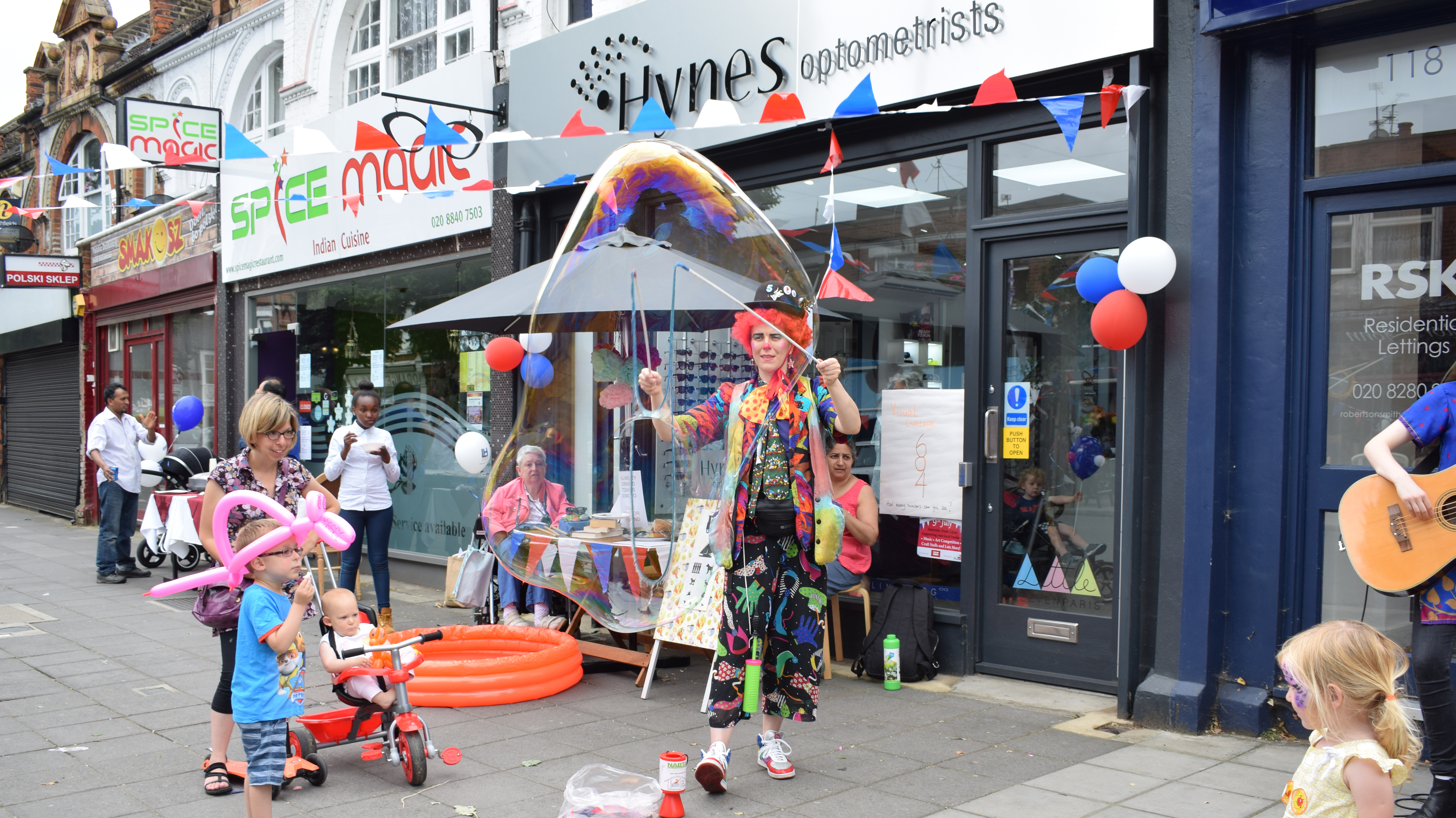 Street event outside Hynes Optometrists with bunting and balloons decorating the practice. A woman and two children are passing by, one child is wearing a balloon hat and the other is in a push chair. In the centre a person dressed as a clown creating large bubbles with two sticks. Behind the clown two women are sitting at either end of a table. On the bottom right a little girl with her face painted and a person playing a guitar.