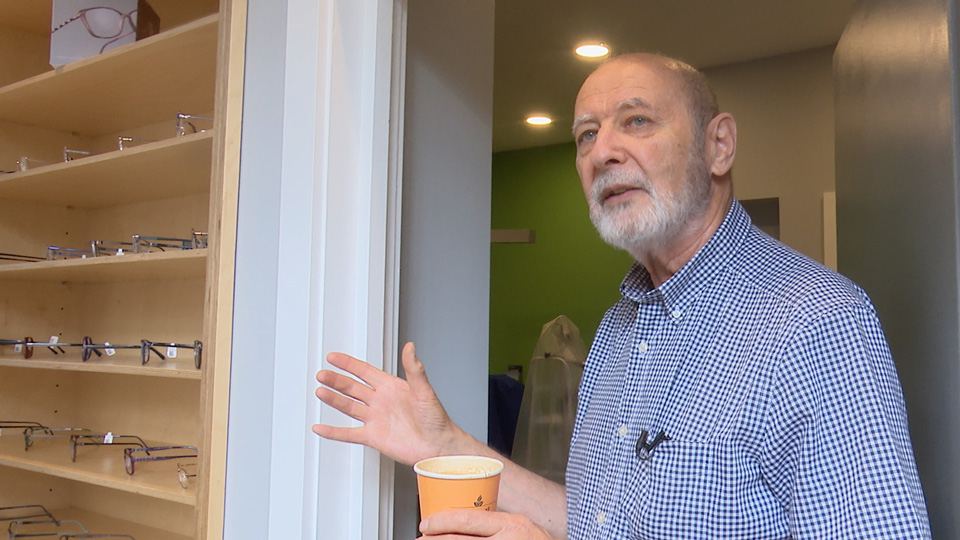 A man in his 70s, wearing a blue and white checked shirt, gesticulates while speaking next to shelves filled with spectacle frames