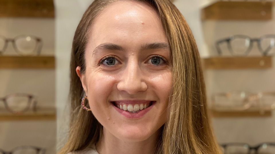 A Caucasian woman with long light brown hair smiles at the camera in front of a display of spectacle frames