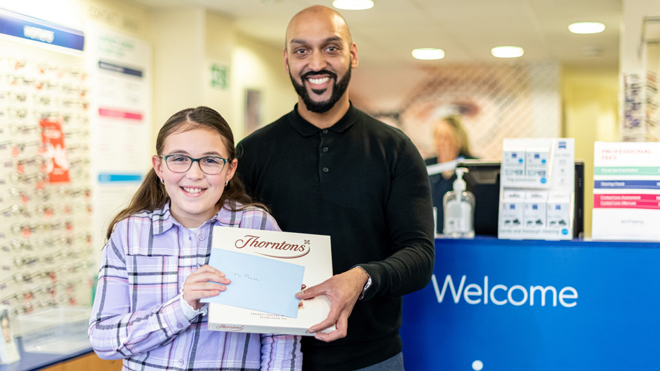 Poppy-May, aged nine, holds a box of chocolates and is smiling standing next to her optometrist Mrudang Patel, also smiling, inside a practice. 