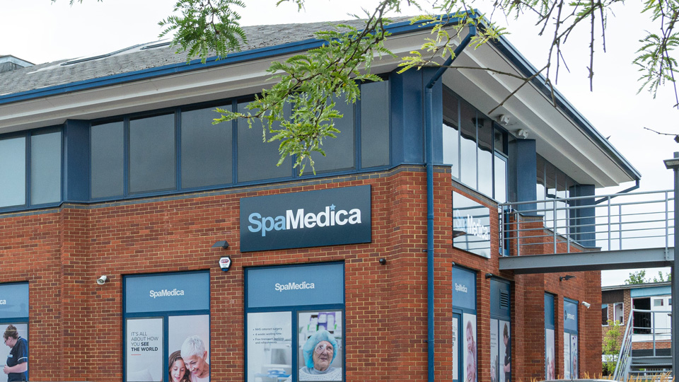 The exterior of a large brick building shows the blue SpaMedica logo, parked cars, and images of patients in covered windows 