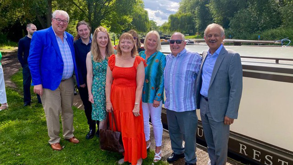 Susan in a red dress standing in the middle with three people standing on both sides of her. The group of people are standing outside on a sunny day in green area, all smiling.