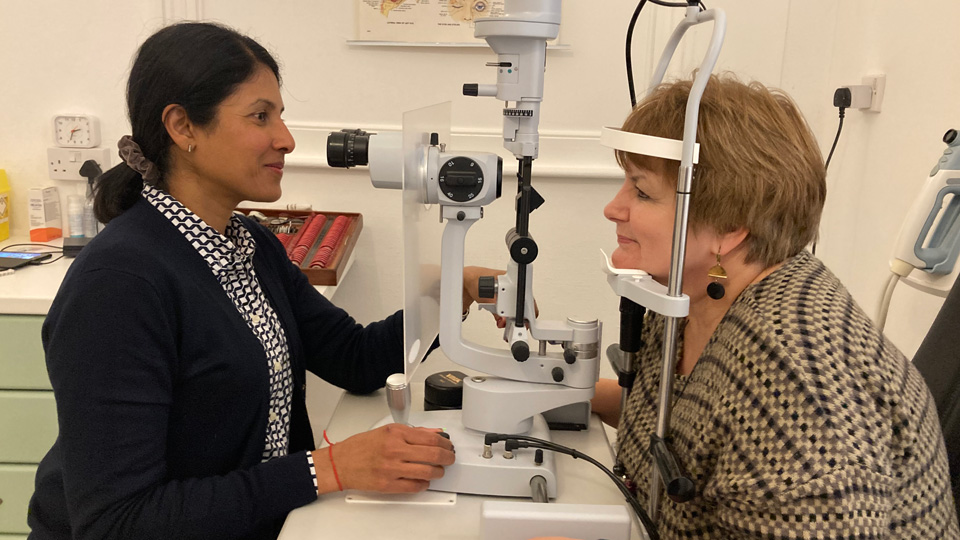 Thanusha Soni wears a blouse and blazer and is testing the eyes of a patient, who is positioned in a slit lamp