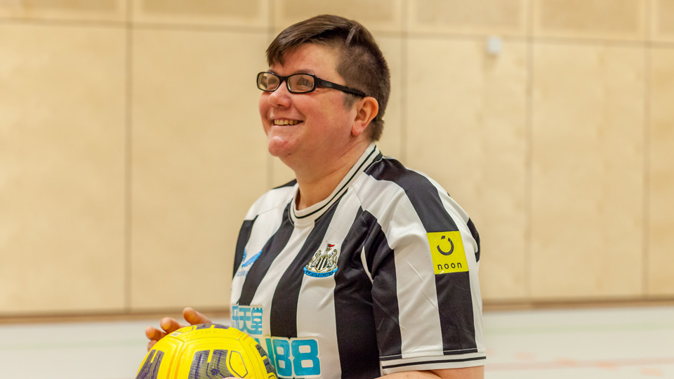 Helen, one of the See Sport Differently supporters, is holding a football and smiling proudly in a sports hall