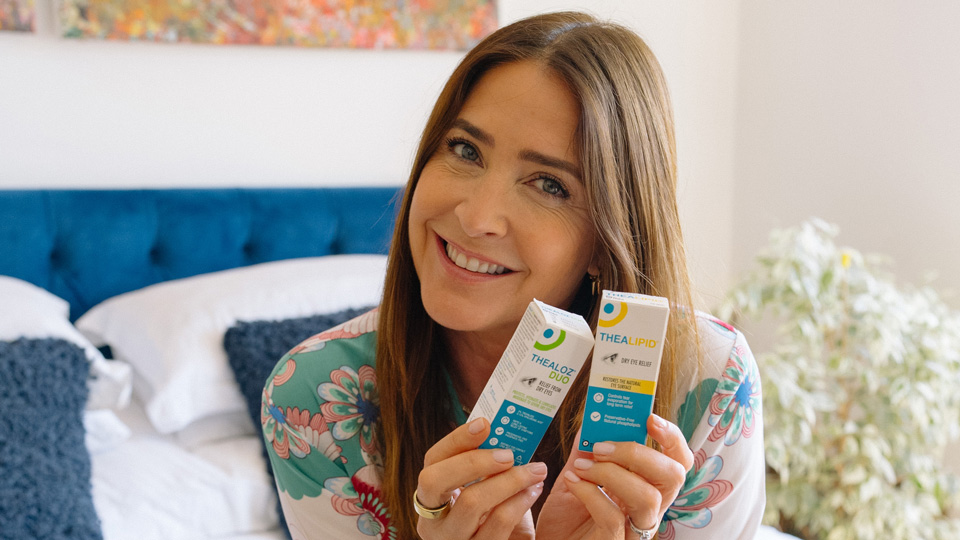 Lisa Snowdon sits on a bed, holding two Théa products up to the camera and smiling
