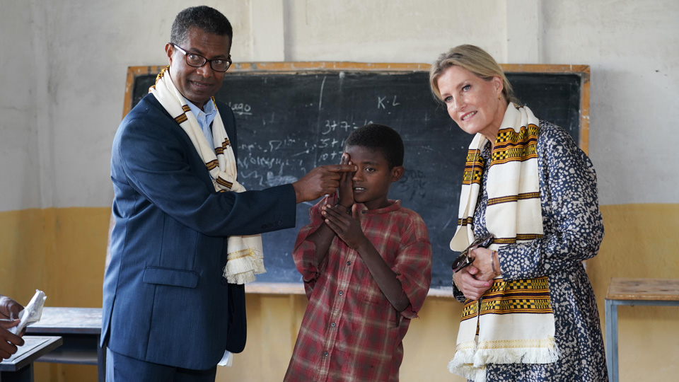 A male teacher dressed in a suit stands with his back to a blackboard and looking across a room, past the camera. He is helping a child beside him to cover one eye for a sight test. The boy, dressed in red flannel, is covering his right eye. Beside him, Her Royal Highness The Duchess of Edinburgh is looking across the room in the same direction as the others, wearing a blue and white patterned dress and a white scarf with a yellow, black and red pattern. 