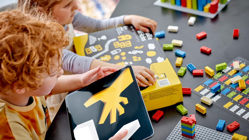 Two children are unpacking the Lego Braille Bricks pack, with lego bricks scattered on a table around them. One holds a tablet showing one of the online activities. 