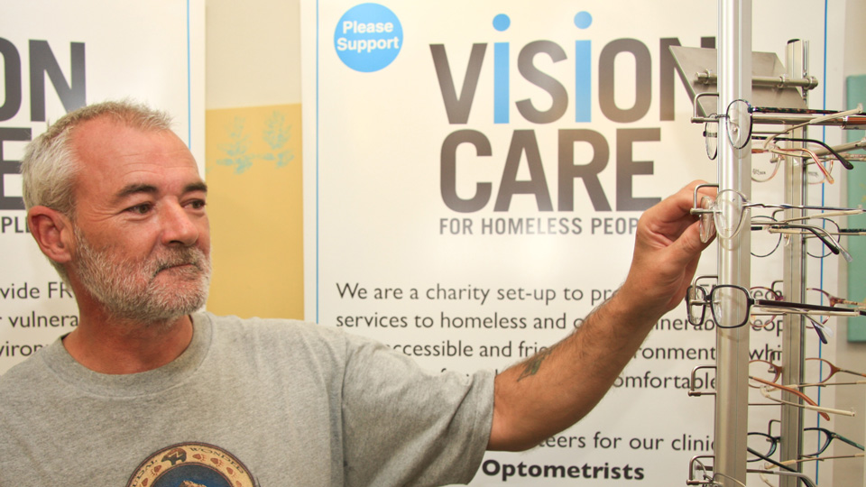 A man is selecting a pair of spectacles from a display. Behind him are banners reading ‘Vision Care for homeless people’