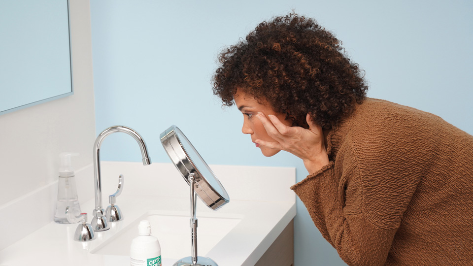 A lady leans over a bathroom counter, looking into a small stand mirror to insert her contact lenses. A bottle of solution and a case for contact lenses are on the side near her