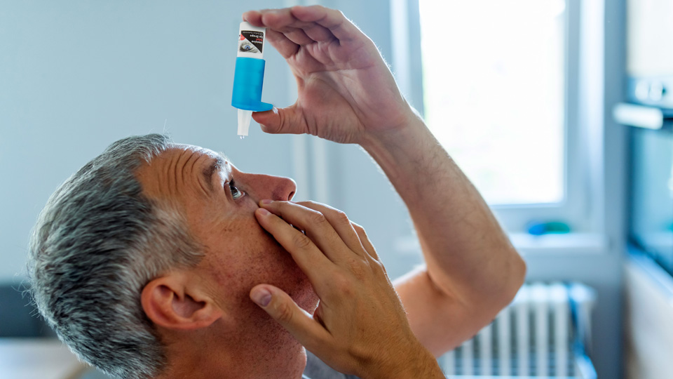 A side profile of a gentleman leaning his head back to apply eye drops. He positions the lower eyelid with one hand and holds the dropper bottle in the air above his eye in the other