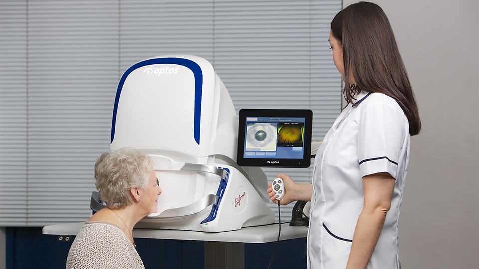 Image shows an OCT machine with an older female patient sat in front of it and a younger female optometrist with brown hair facing towards the OCT and away from the camera 