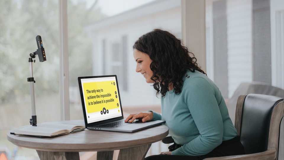 A woman is seated at a table. The Orcam device is positioned on a stand above an opened book. Beside her is a laptop