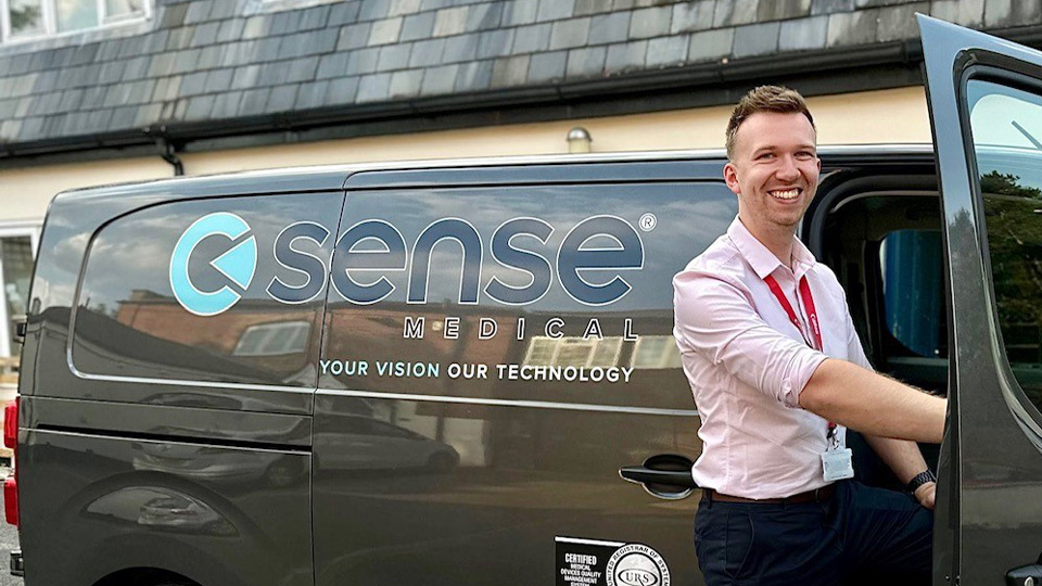 Joe Blamire is photographed by the open door of a black van with ‘Sense Medical’ printed along the side. He wears a shirt with sleeves rolled to his elbows and smiles wide at the camera.