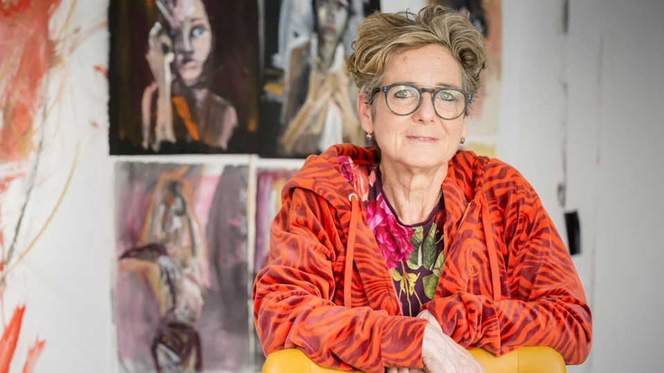 Rachel Gadsden wears grey aviator-style glasses, a bright orange tiger-striped hoodie and stands behind a mustard yellow chair. On the chair is a painted portrait, and more portraits hang on the wall behind her