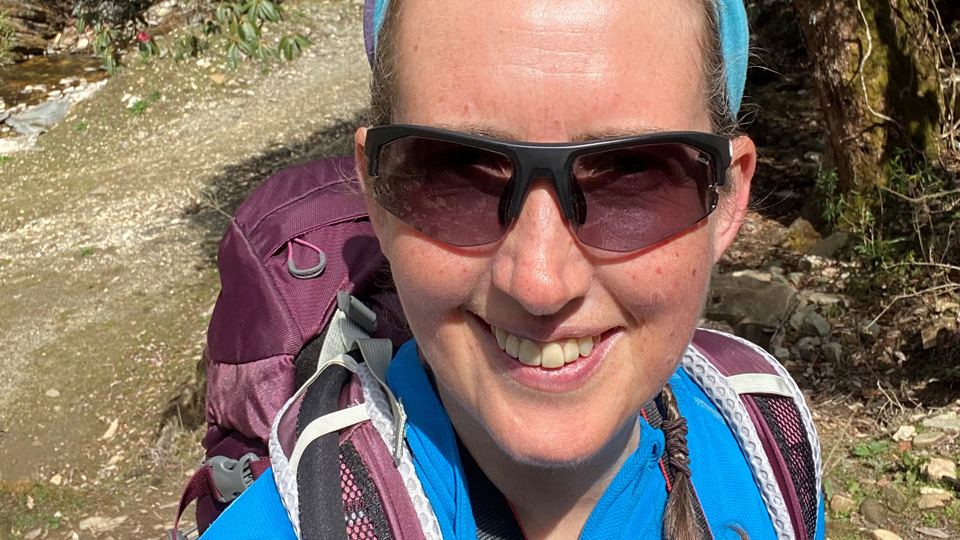 Elaine is standing in a wooded area with chalky, sloping ground behind her. The selfie shows her wearing a blue walking jacket, mauve purple backpack, and light blue fabric headband. Her hair is tied back in a plait and she wears a pair of Bollé sunglasses. The wrap around frame has lenses that appear to be a purple tint. 