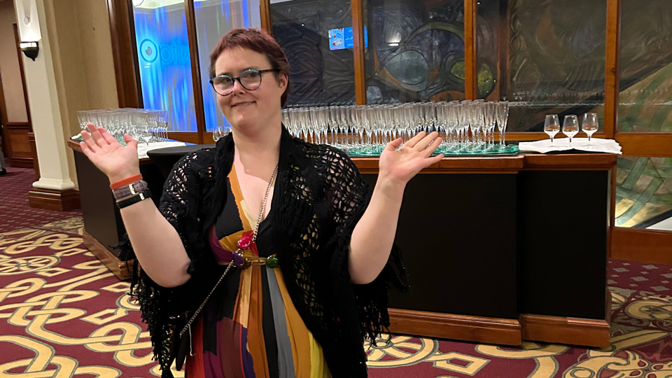 Grace stands in the lobby of the Optix conference, raising her arms and smiling for the camera. She wears blue and purple spectacles and a long colourful dress. 