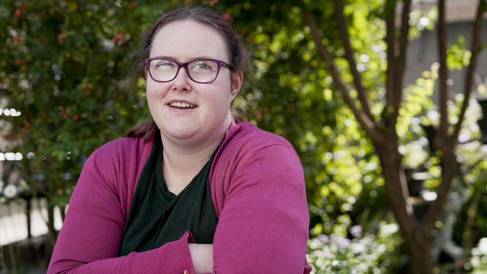 Grace smiles to the camera, wearing dark purple spectacles, a black top and purple cardigan 