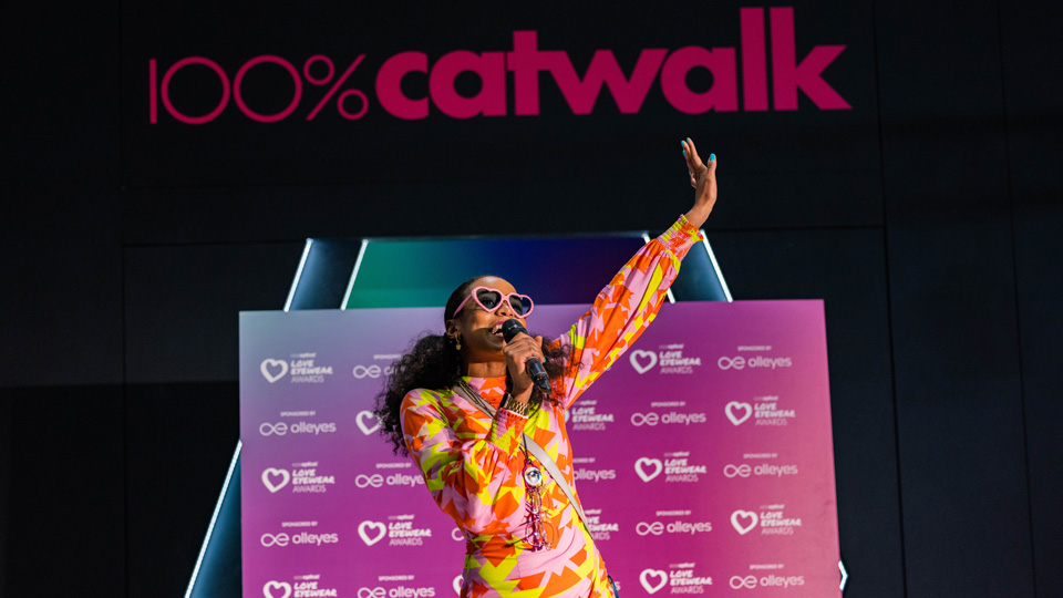 A woman wears oversized pink heart sunglasses and a bright patterned dress of oranges, pinks and yellows. She is speaking excitedly into a microphone in front of signs reading 100% Catwalk  