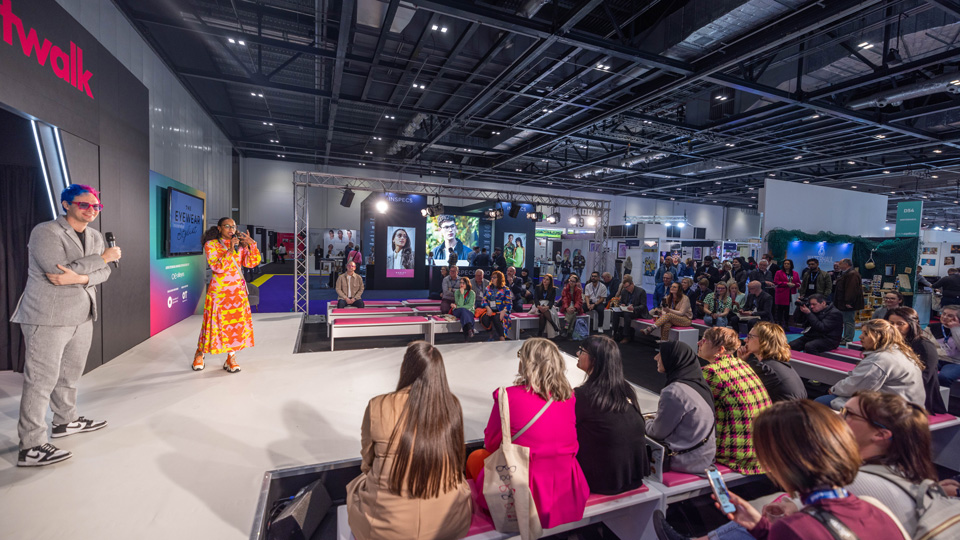 People are gathered on pink bench seating around a catwalk stage looking towards the two hosts – one wearing an orange and yellow patterned dress, and the other in a light grey suit with hair dyed blue and pink