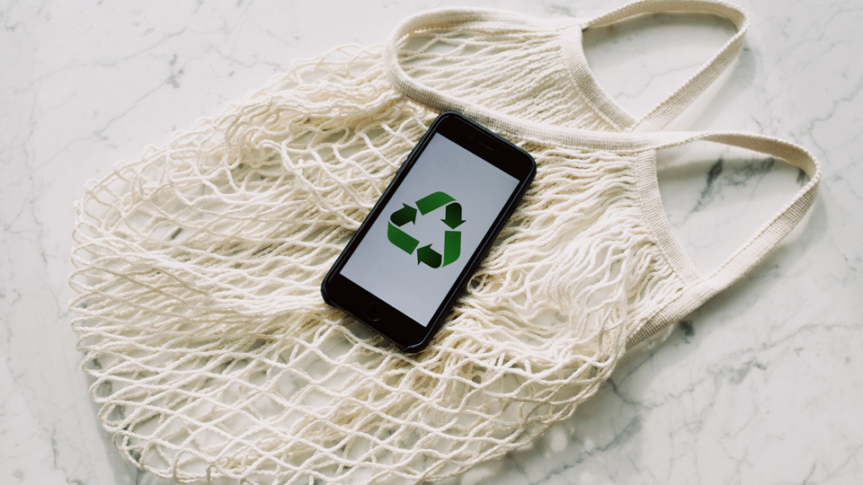 A smartphone displays a white screen with the recycling icon of three arrows pointing in a rounded triangle. The phone lies against a white net bag.