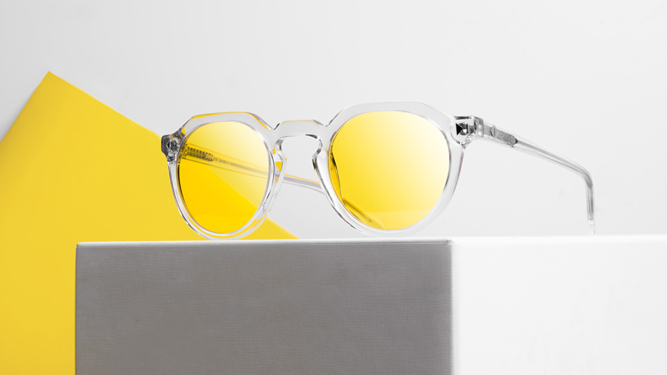 A slightly squared clear frame is positioned on a grey cube. The lenses of the frame are a bright yellow gradient