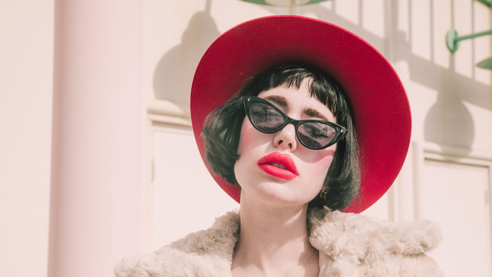 A model wears a pair of black sunglasses in a sharp cat’s eye shape. The model has very pink blusher, bright red-pink lipstick and a wide-brimmed red hat. She wears a white fluffy jacket and stands against a white background