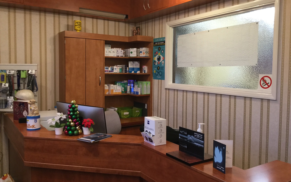 A heavy wooden reception desk fills the frame, with lens wipes and Christmas decorations. Behind it is a large wooden cabinet with a range of eye drops and treatments. The walls have a cream striped wallpaper and large internal window with papers tacked to the other side