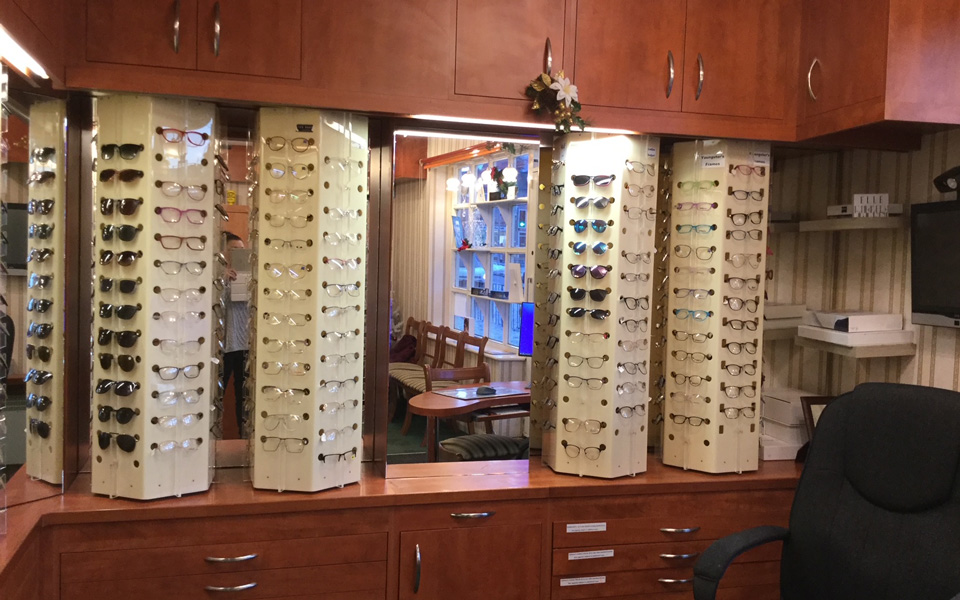 The dispensing area has large wooden cabinets and drawers. Between them are column stands of eyewear frames. A mirror reflects the large window and a row of wooden chairs with striped seats.