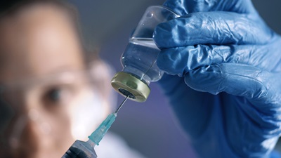 Image shows a blue gloved hand injecting a clear substance into a vial 