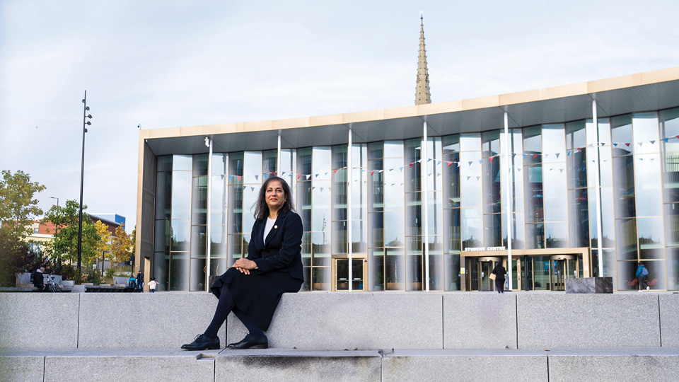 A woman in a dark suit sits on stone steps in the distance. Behind her is the glass front of a tall, modern building with a spire rising above the building