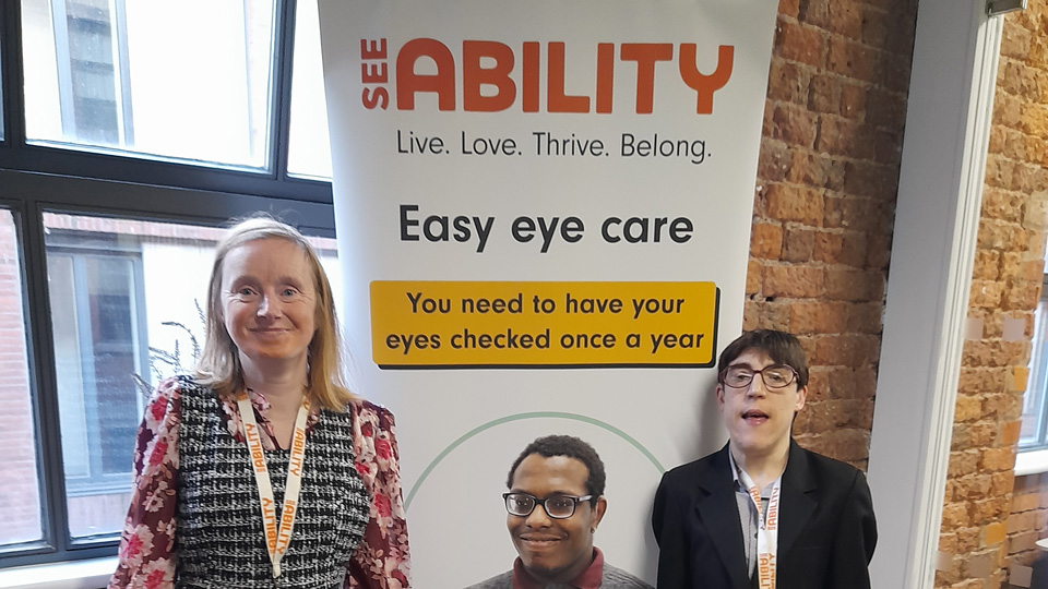 Joanne and Rebecca, both dressed smartly and wearing lanyards, stand in front of a banner for the SeeAbility, with a title on the Easy Eye Care pathway