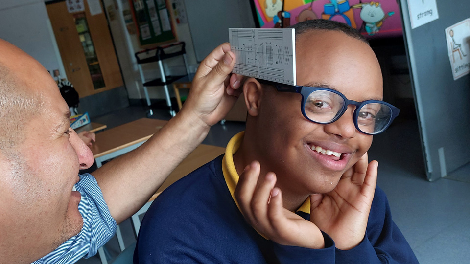 A man holds a measuring ruler up to the temple of a boy wearing glasses and smiling
