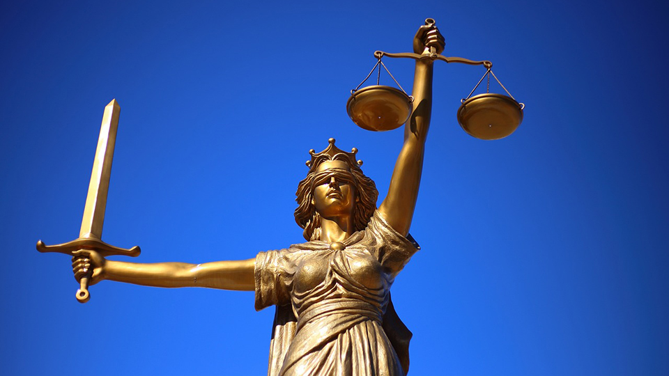 A gold statue of a woman in a robe stands in front of a blue background. In one arm she holds a sword. In the other arm she holds a set of scales