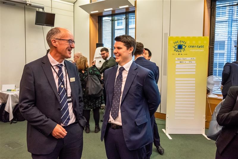 Thom Renwick and Ben Burton are laughing in a room in parliament in front of a yellow sign for Westminster Eye Health Day 2022