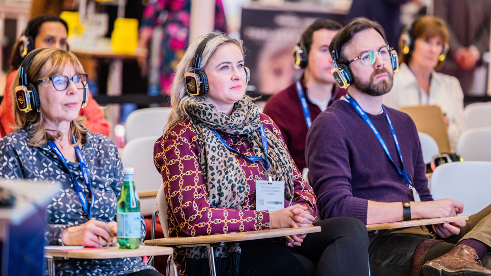 An audience of professionals are engaging in a lecture. Three individuals are in frame, each sat at desks and wearing headphones. They look off to the right and a presentation is reflecting in the glasses of two of the delegates. They all wear smart causal, patterned clothing. More audience members are behind them, but blurred. 