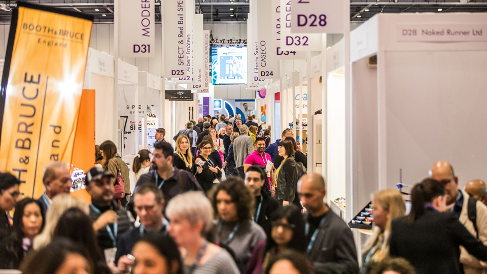 A bustling crowd fills the corridor between stands in the 100% Optical exhibition hall