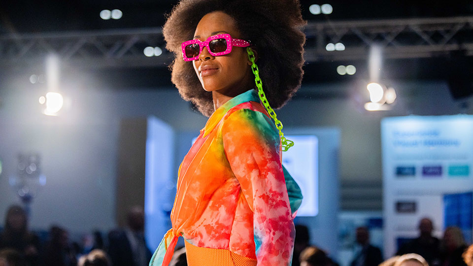 A woman poses on a catwalk wearing a bright top with a tie-dye style of oranges, yellow, pink and blues. She wears chunky bright pink rectangle sunglasses, with neon green glasses chains 