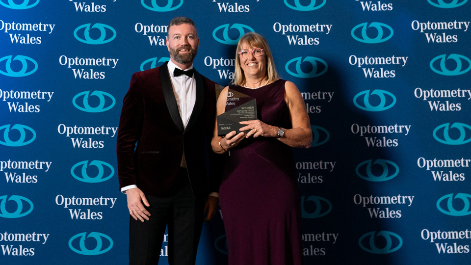 Helen stands in front of a wall with the Optometry Wales logo, holding a trophy and wearing a floor-length wine-red dress
