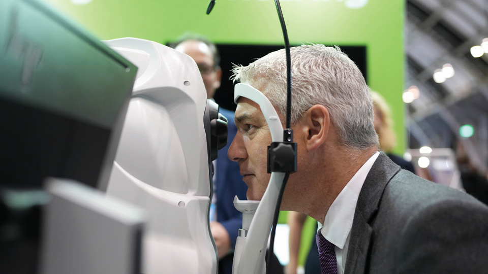 Side view of Steve Barclay, the former health secretary, who is looking into a white OCT machine. In the background is lime green Specsavers signage 
