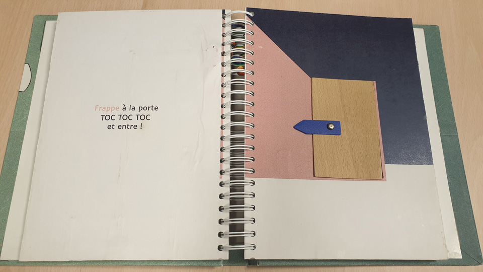 A book is open on the table. A door is pictured on the page with a small metal button that forms the handle. 