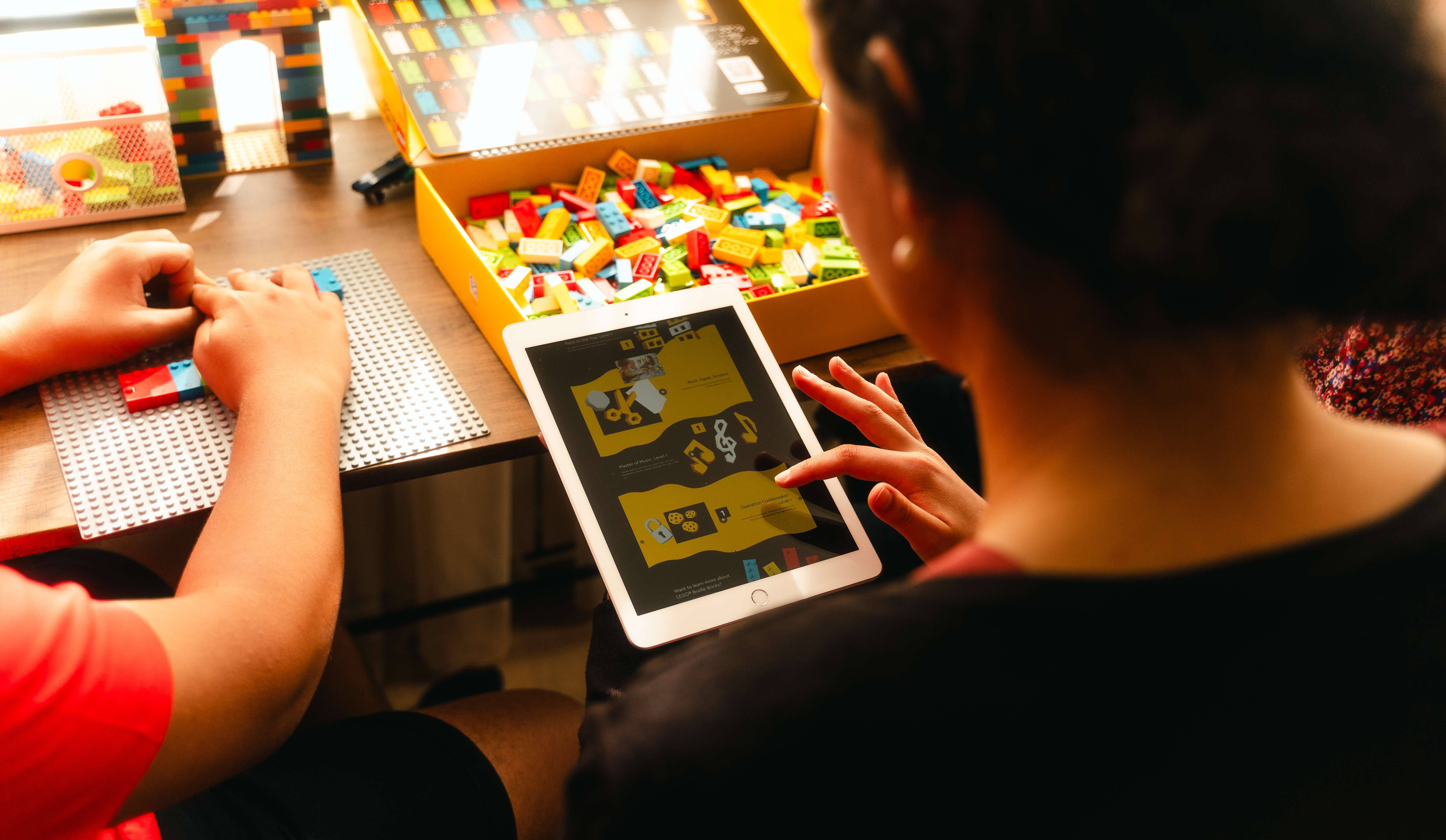 A woman looks at a tablet screen displaying one of the Lego Braille Bricks online PlayStarter activities. An open box of Lego Braille Bricks on a desk before her, hands build with Lego Braille Bricks on a baseplate on the desk beside the box.