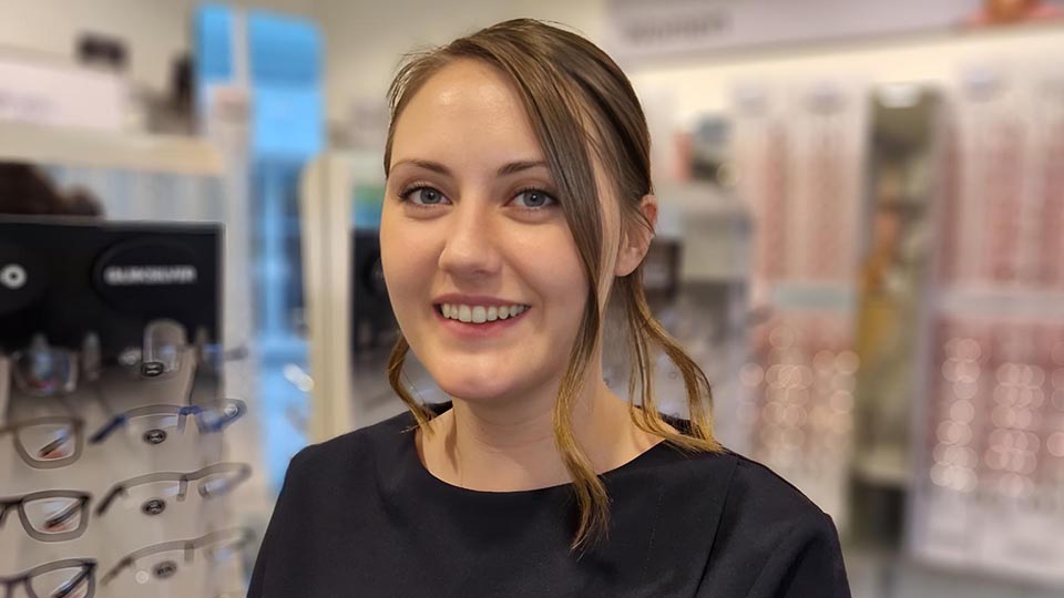 Cat Cooper is wearing a navy blue Specsavers uniform and is smiling in front of a display of trial frames, with her brown hair in a ponytail 