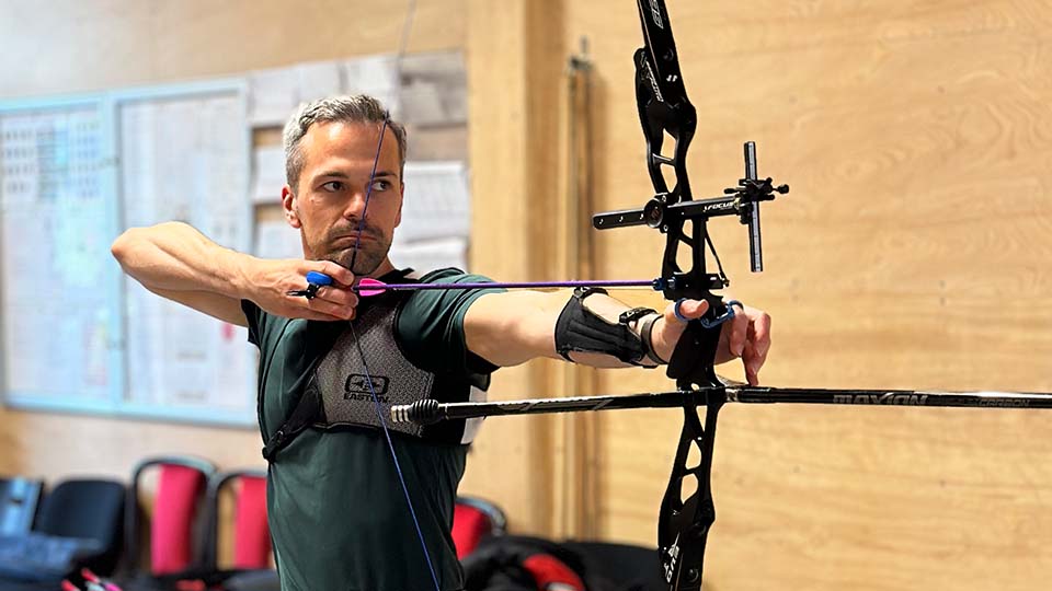 Nick Clapham is wearing blue jeans and a green t-shirt and is holding a large bow and arrow inside a wood panelled room 