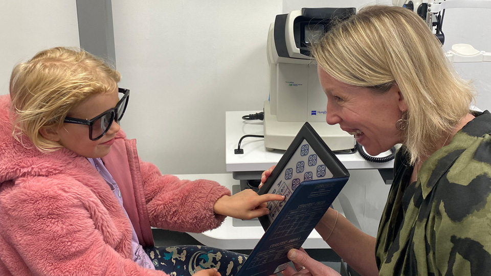 Young child wearing a pink jacket and black sunglasses in the testing room. She is sitting opposite an optometrist with short blonde hair.