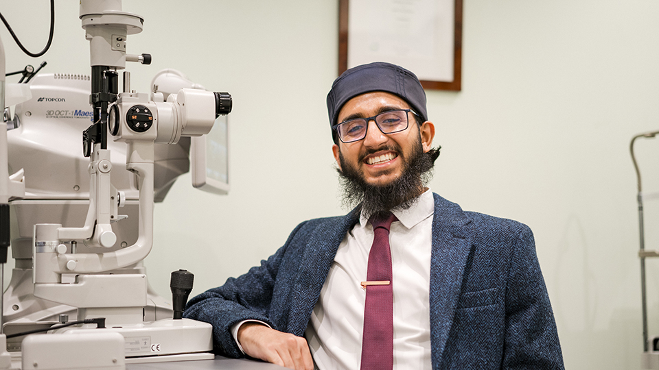 Habeeb is sitting at a desk next to a large piece of optical equipment and is smiling