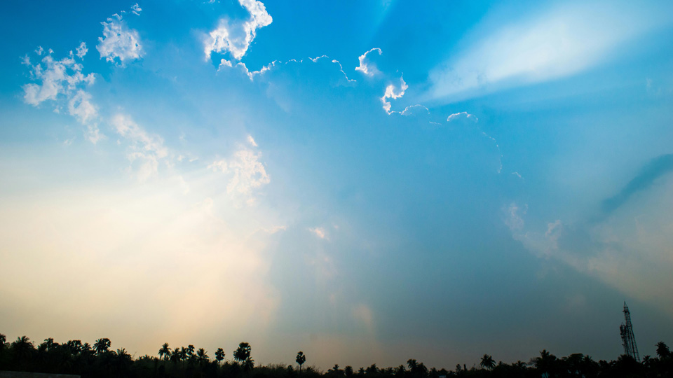 The sunlight flares behind fluffy white clouds on a blue sky, with a silhouette of a hill framed along the bottom of the frame