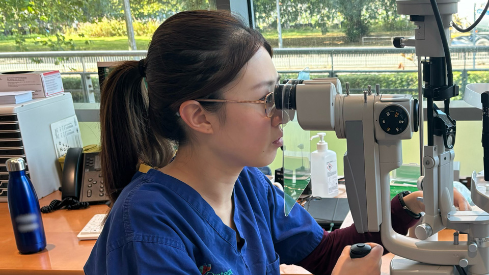 Bonnie is operating a device in a clinical setting. She wears dark blue scrubs with the Optegra logo and her hair is pulled back into a ponytail