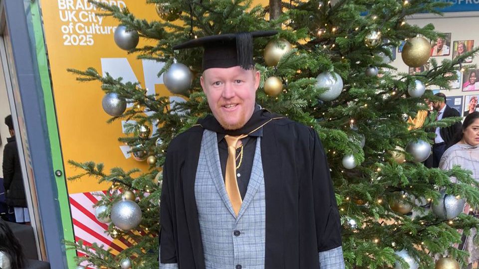 Wearing a graduation gown, Daniel Morgans stands by a sign for the University of Bradford